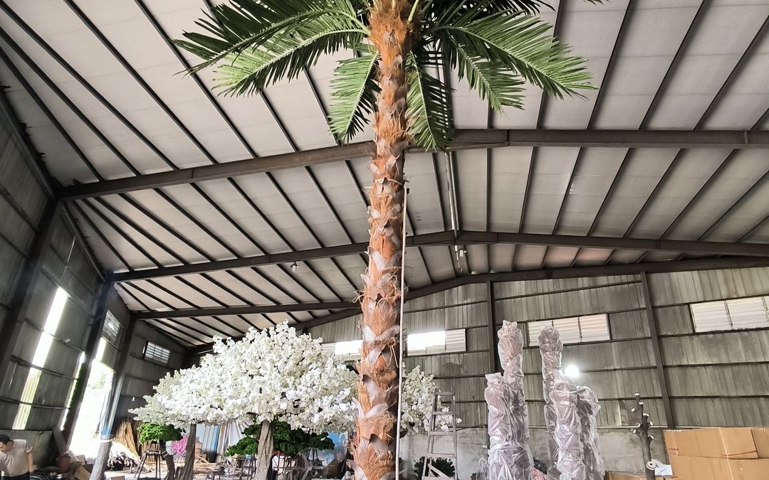 Tall artificial date palm photographed looking up towards the workshop roof, with a white blossom tree and wrapped trunk stock nearby