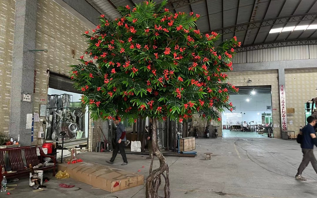 Artificial flowering tree with red blossoms and green canopy standing inside a factory workshop, a worker walking past