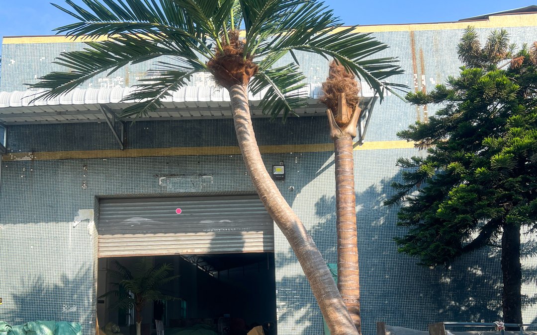 Two curved palm trunks with brown finish and green frond crowns standing outside the factory gate