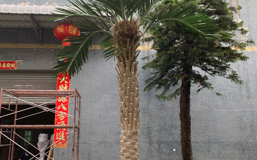 Two artificial palms standing outside a workshop entrance, trunk moulding and crown fibre visible