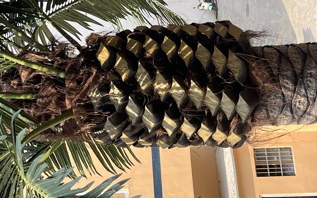 Close-up of a molded artificial palm trunk with diamond leaf-scar pattern and fibre tufts, staged outdoors against a factory wall