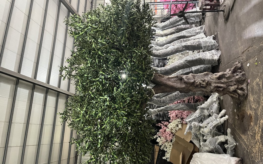 A finished artificial tree standing on the workshop floor with a row of film-wrapped trunks staged behind it