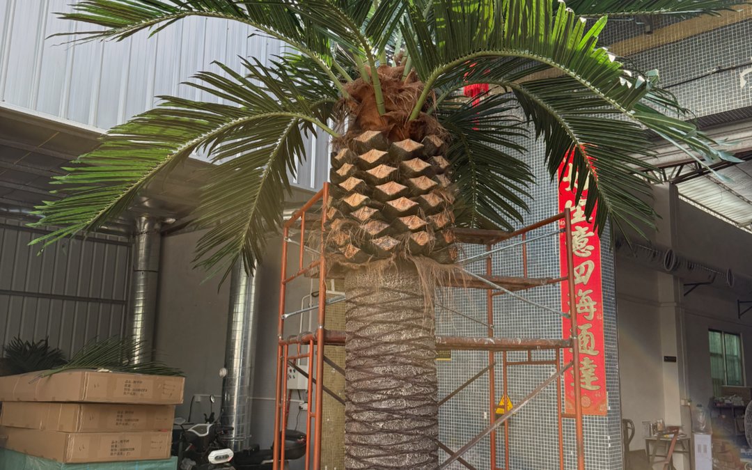 An artificial palm under scaffolding at the factory, showing the diamond-pattern trunk and frond crown