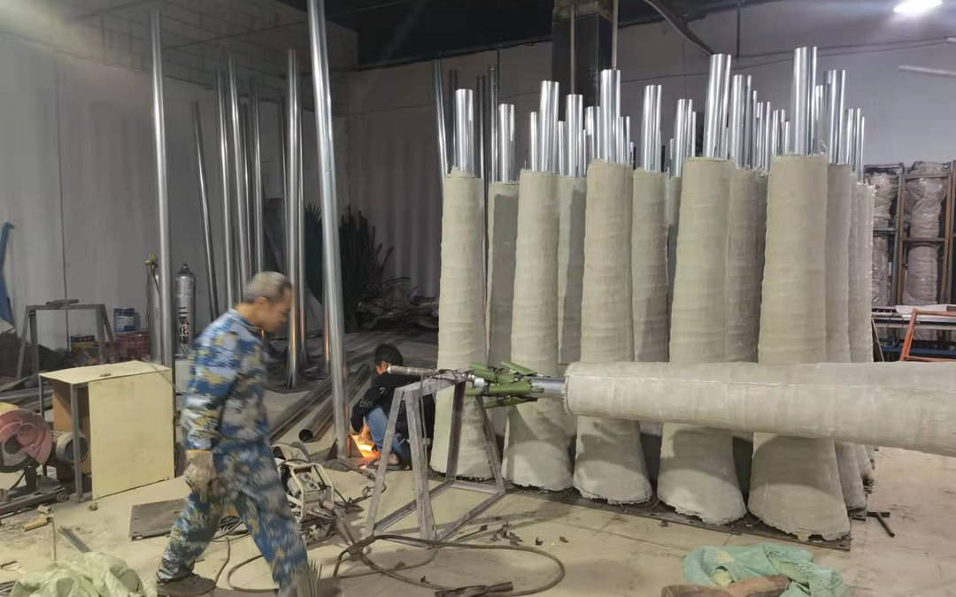 Rows of upright trunk cores wrapped in fabric sleeves with steel tubes protruding, in a production workshop