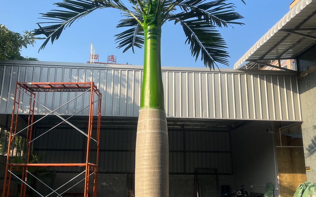 A tall artificial palm standing outdoors in a factory yard against corrugated metal buildings