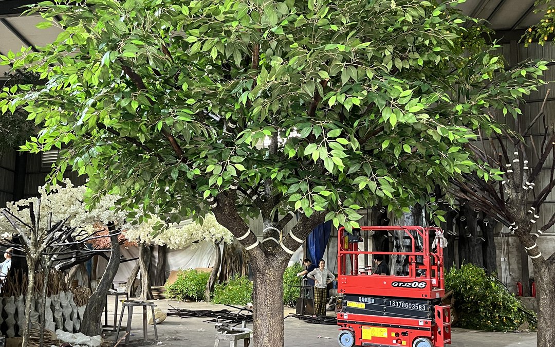 Large artificial ficus tree with banded trunk standing beside a scissor lift in the production yard