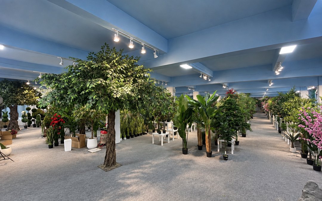 Rows of artificial trees and planters displayed in a factory showroom hall, with a large broadleaf tree in the foreground