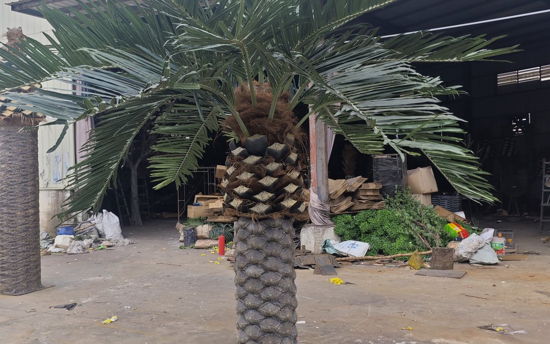 Artificial palm dry-assembled in a factory yard, molded diamond-pattern trunk and fibre collar at the crown base, fronds staged flat behind it
