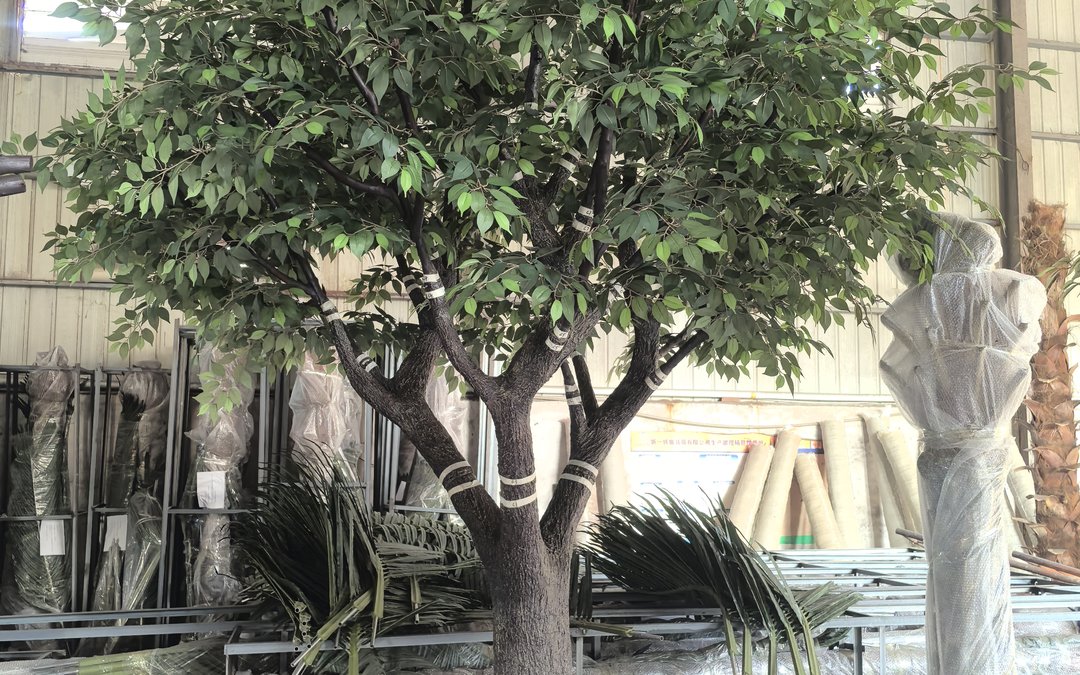 Evergreen broadleaf-style artificial tree indoors with white banding wrapped at trunk and branch junctions, surrounded by semi-finished trees and steel frames
