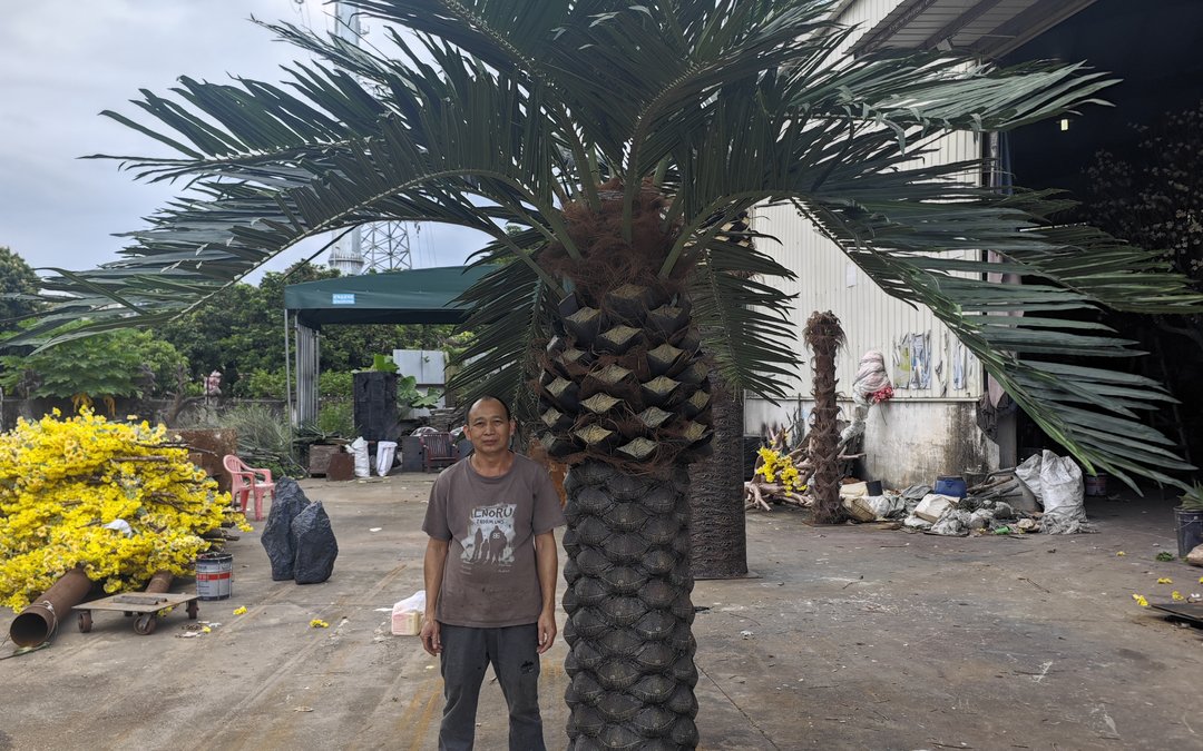 Full-height artificial palm with feather fronds and scaled trunk standing outdoors, with a person beside it for scale reference