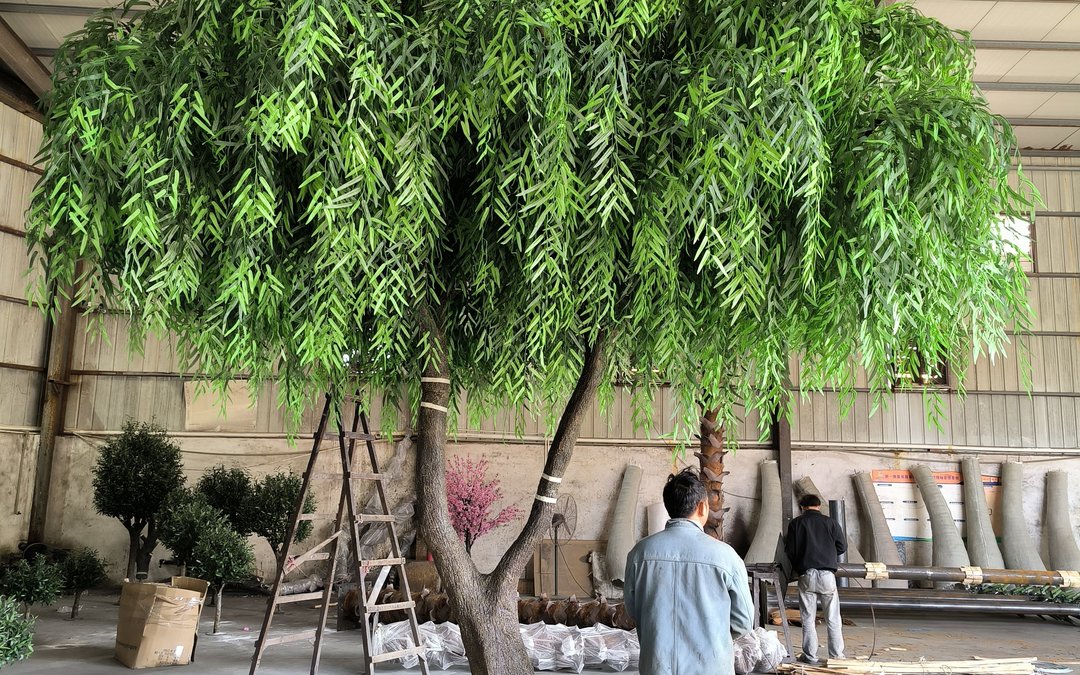 Large artificial weeping willow with dense trailing foliage indoors, two workers standing beneath it for scale, semi-finished trunks and steel frames behind