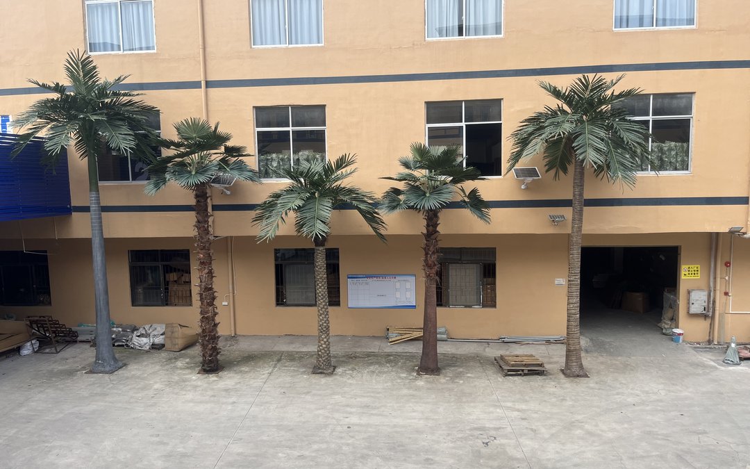Five artificial palm trees of varying height and canopy shape standing in a row on the forecourt of a factory building
