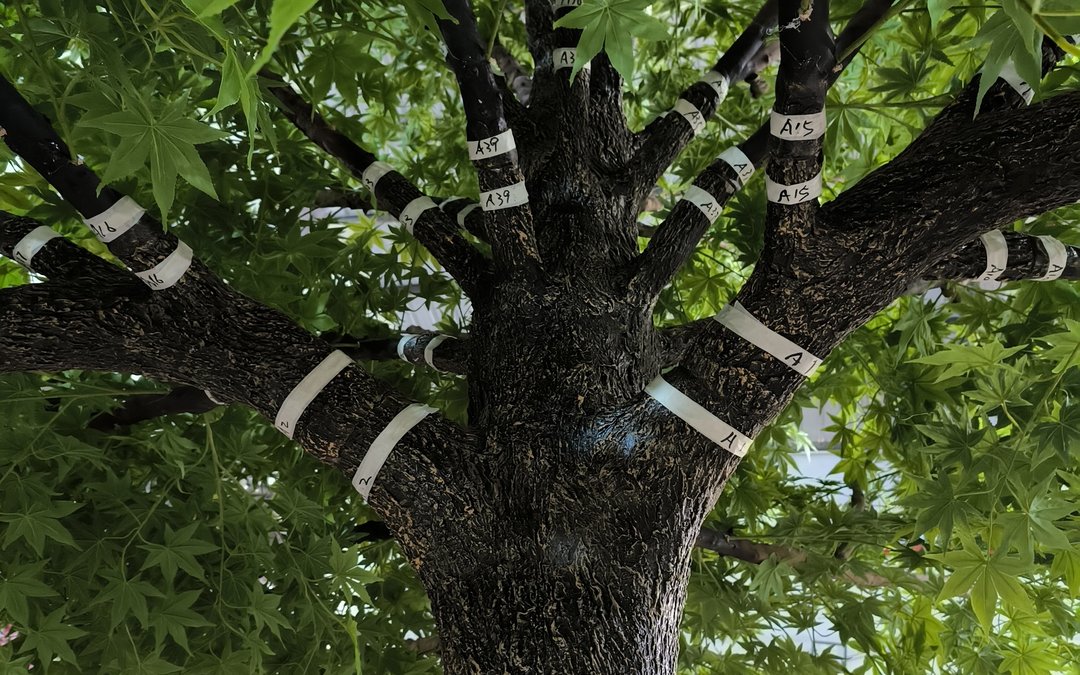 Branch arms carrying hand-lettered assembly tags on a dark trunk, maple foliage behind
