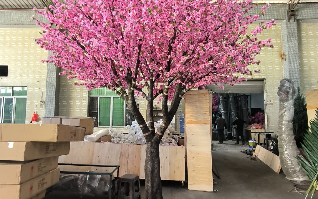 Pink blossom tree standing in the factory yard among plywood crates and cartons, with wrapped trunk lengths beside the loading bay