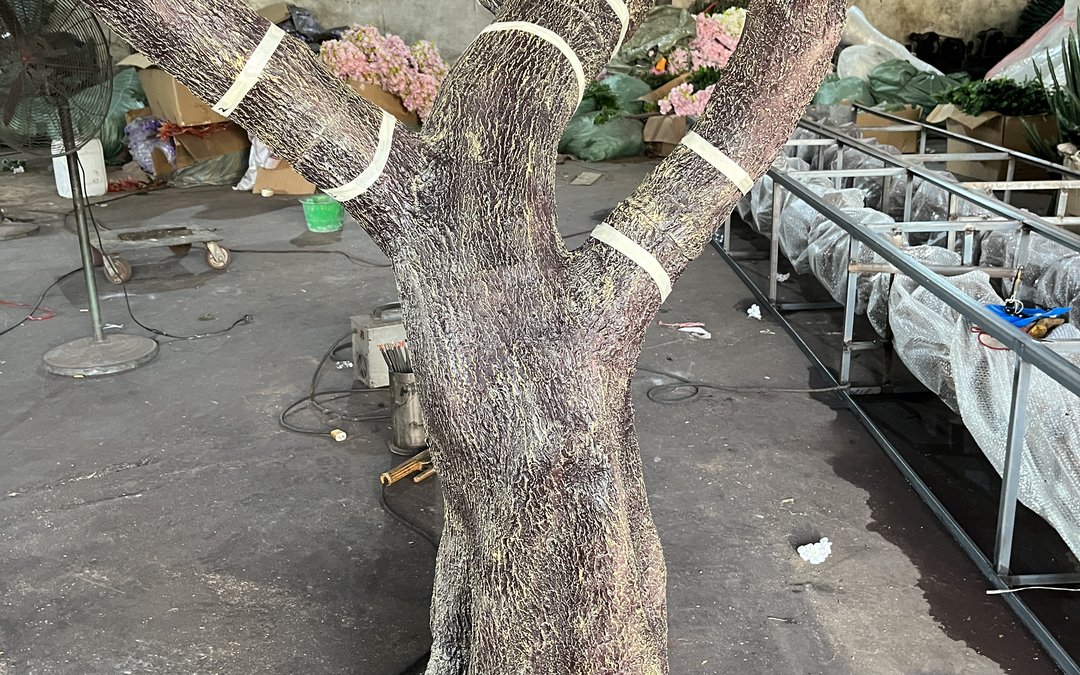Large artificial tree trunk on the workshop floor with protective bands at the branch junctions and wrapped components on racks behind