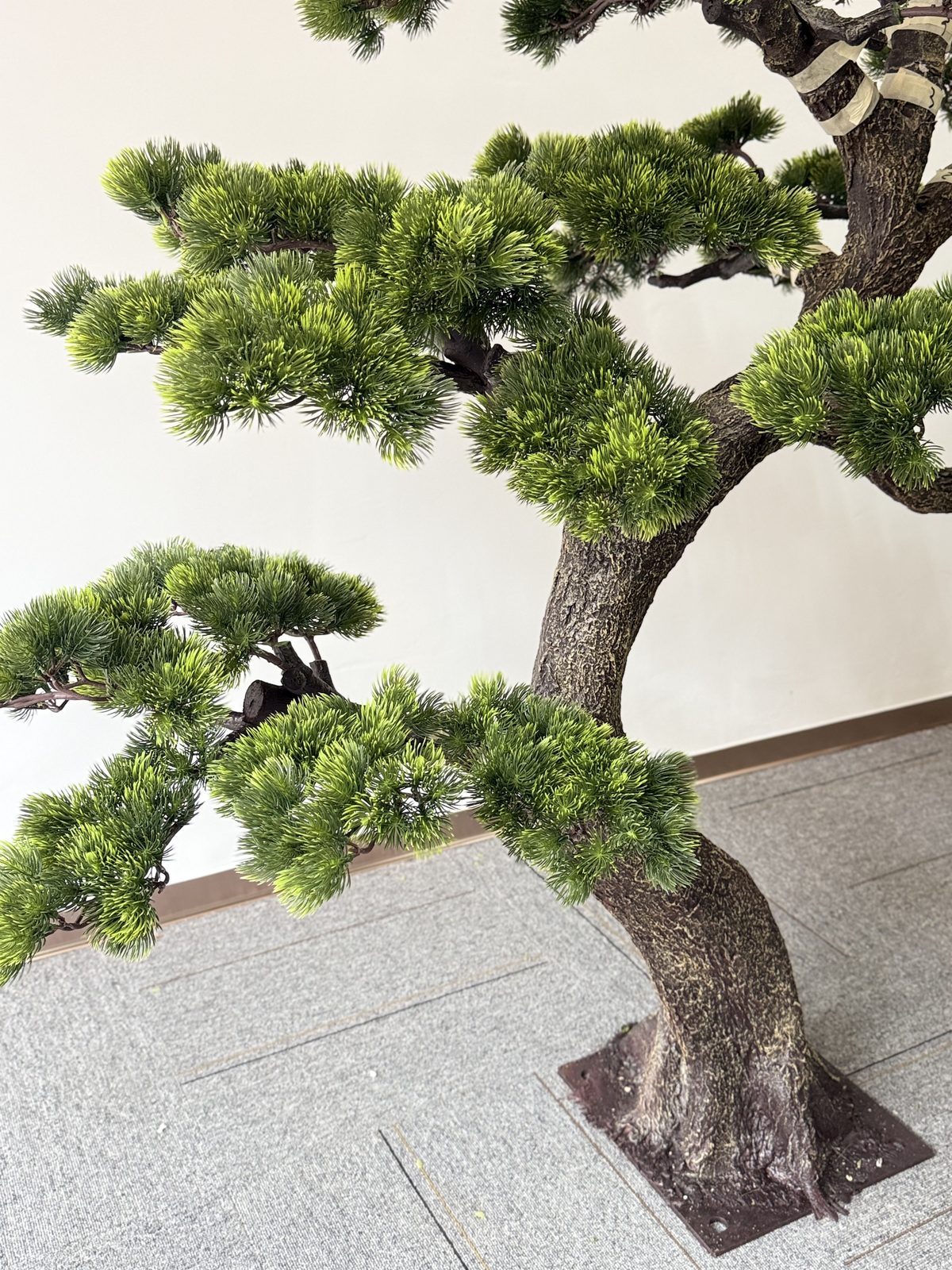 Bonsai-style artificial pine with fine needle pads against a plain interior wall